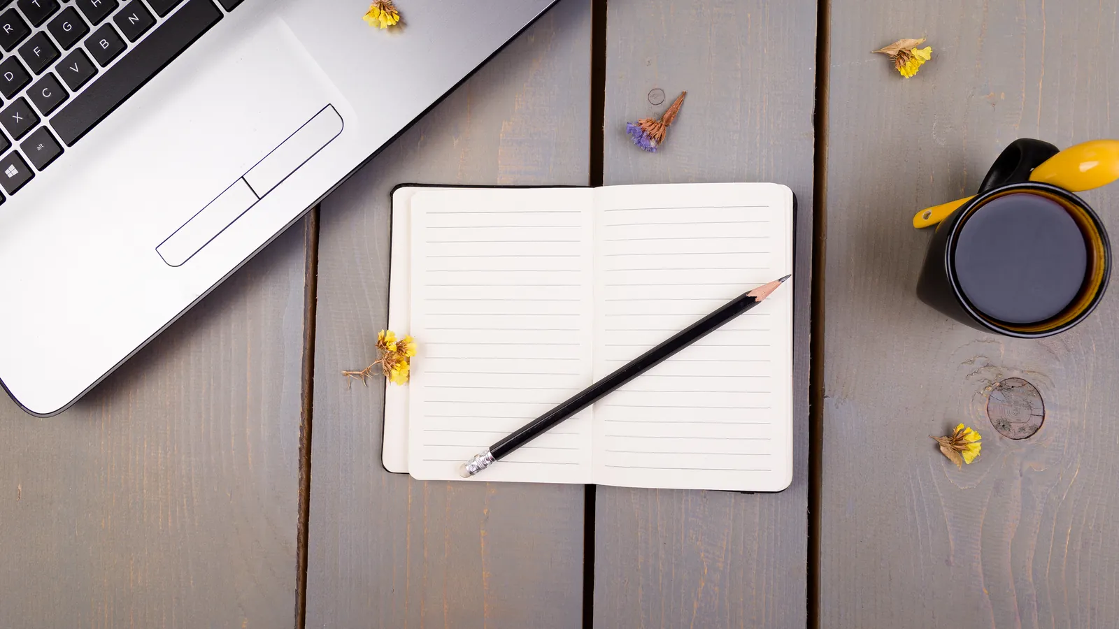Bureau en bois avec un ordinateur portable, un carnet ouvert, un crayon, une tasse et des fleurs jaunes
