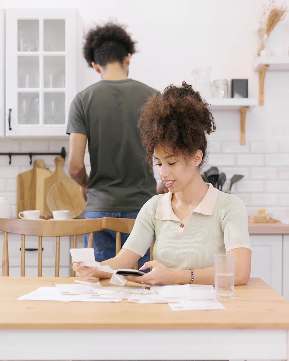 Une femme vérifie des factures sur la table de la cuisine pendant qu’un homme se tient de dos près du plan de travail